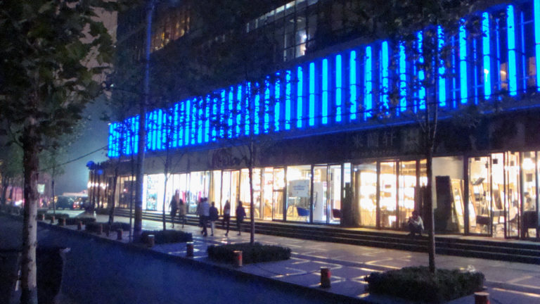 Wide street view of a modern Beijing retail facade lit by vertical blue LED bars above bright storefront windows with pedestrians and trees along the sidewalk.