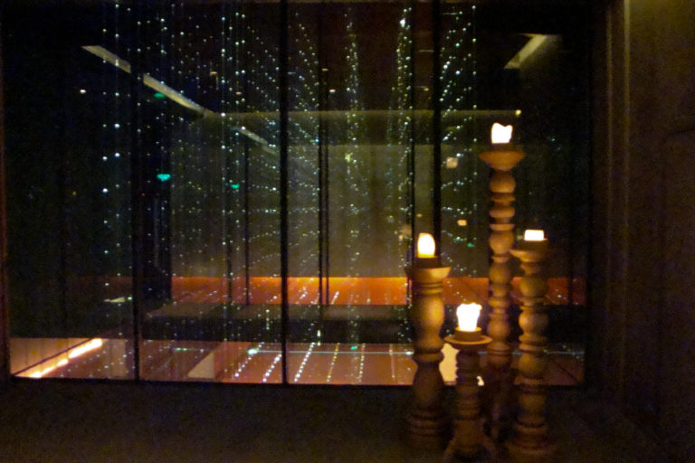 Tall sculptural candleholders of different heights glowing beside a reflective glass wall dotted with tiny points of light.