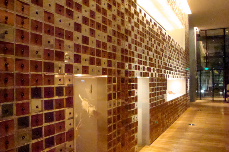 Corridor with a long wall of small wooden apothecary-style drawers and illuminated display niches leading toward a glass doorway.