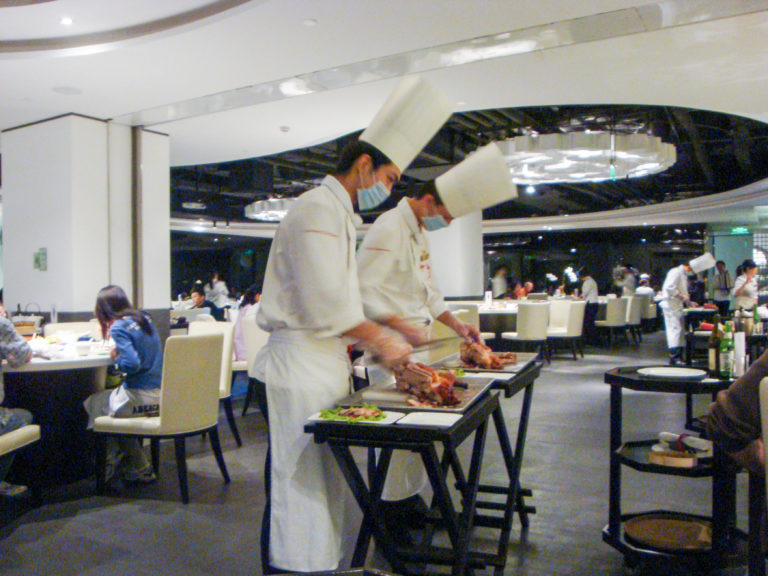 Two chefs in white uniforms carve roast Peking duck on portable tables beside seated diners in a Beijing restaurant.