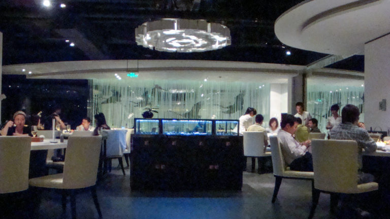 Wide view of a modern upscale Beijing restaurant dining room with pale chairs, white tables, large circular ceiling lights, and a glass-sided central installation surrounded by diners.