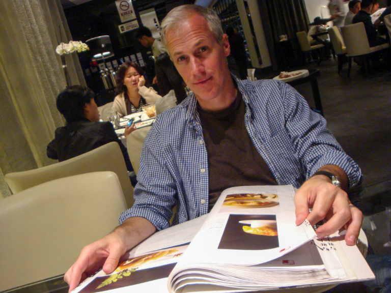 Gray‑haired traveler holding open an illustrated menu at a restaurant table during an evening meal, with other diners visible in the background.