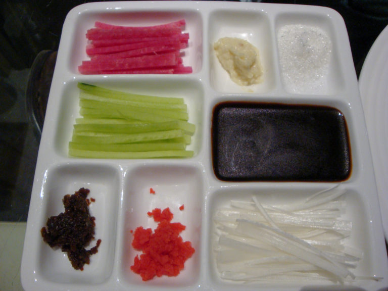 Overhead view of a compartmented white dish holding cucumber, scallion, dark sauce, sugar, and other condiments for Peking duck.
