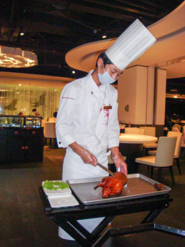 Chef in white uniform and mask carving a glossy whole Peking duck on a board in a restaurant dining room.
