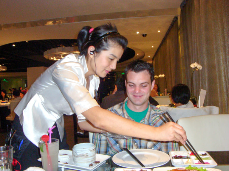 Female server in a light uniform leans across a table to serve a seated diner in an upscale Beijing restaurant.