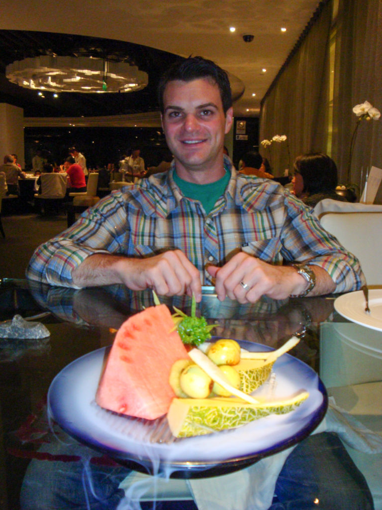 Traveler in a plaid shirt seated at a glass table in an upscale Beijing restaurant, with a decorative fruit plate featuring a large watermelon wedge in the foreground.