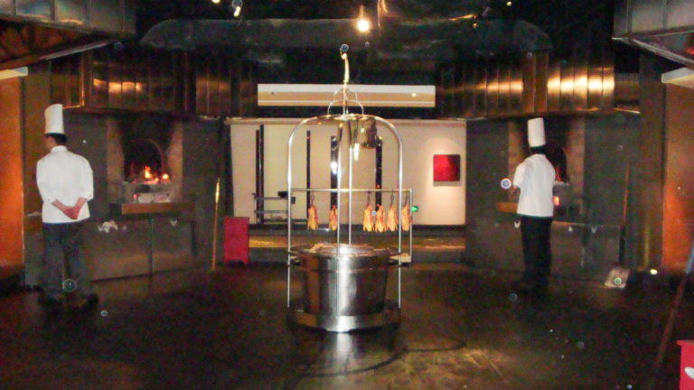 Two chefs in white uniforms stand beside a stainless-steel cylindrical roasting apparatus in the open kitchen of a modern Beijing restaurant.