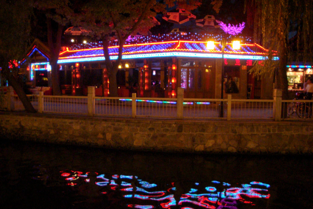 Traditional-style waterfront bar in Beijing outlined with multicolored lights, reflected in the calm canal water at night.