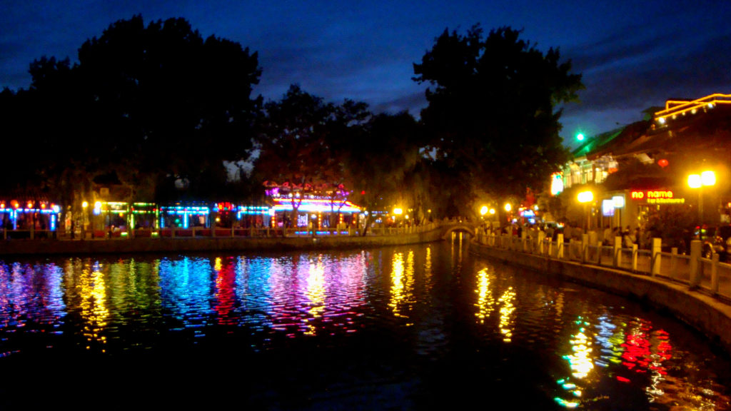 Night view of illuminated waterfront venues along a narrow canal or lake in Beijing’s Shichahai–Houhai area, with colorful lights reflecting on the dark water and trees framing the scene.