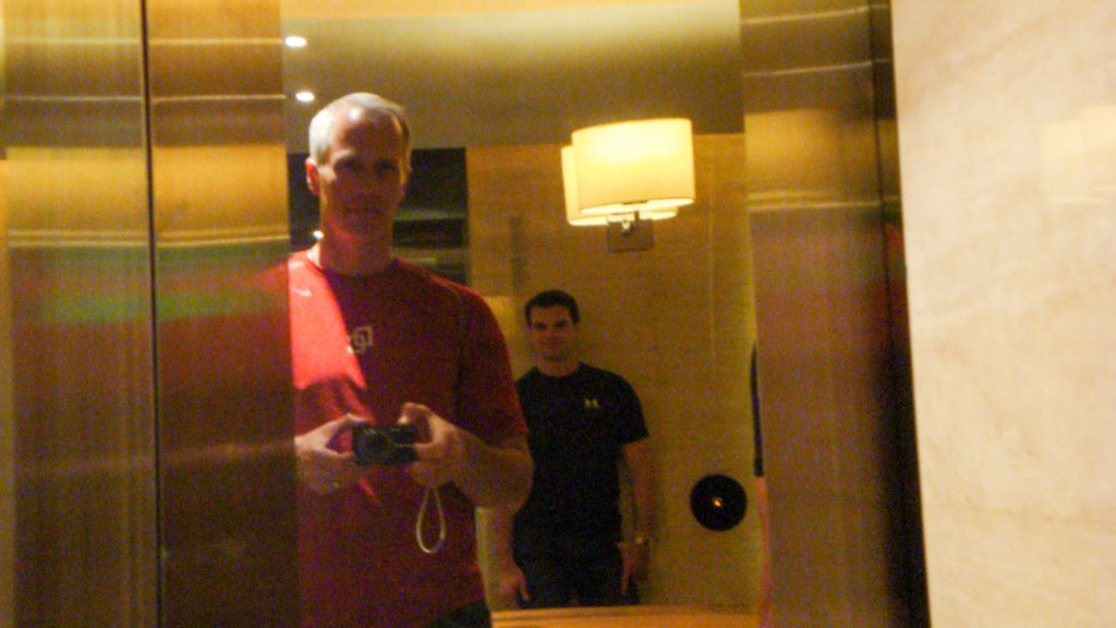 Two travelers take an informal mirror self-portrait in a softly lit Beijing hotel room, with one holding a camera at chest height and the other standing behind.