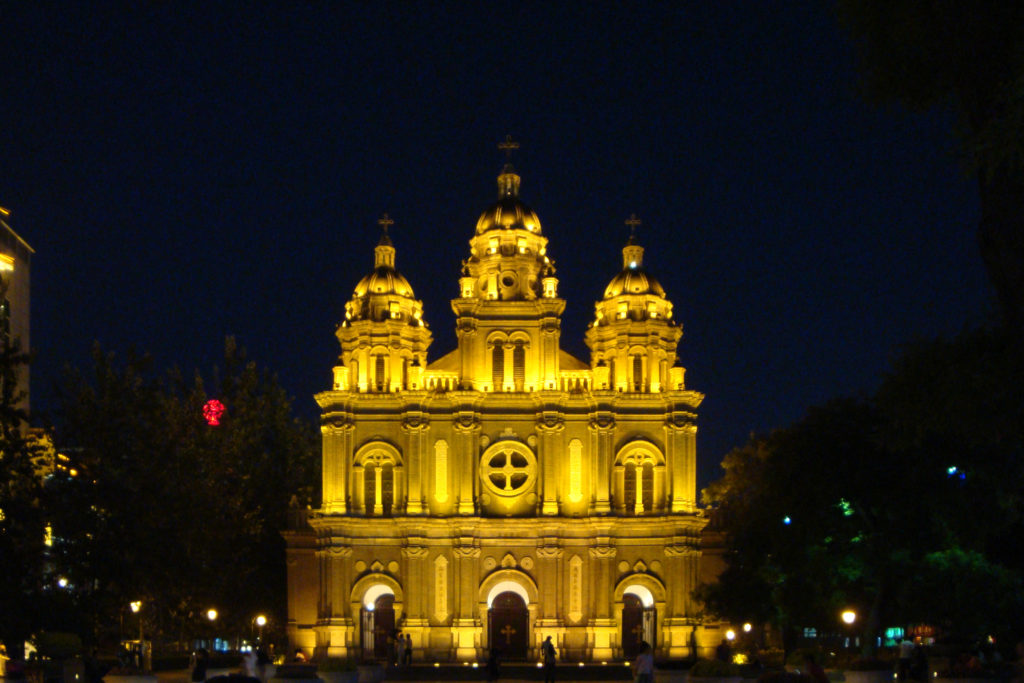 Distant night view of the floodlit facade and three towers of St. Joseph’s Church in Wangfujing, seen across a dark plaza with small visitors and trees.