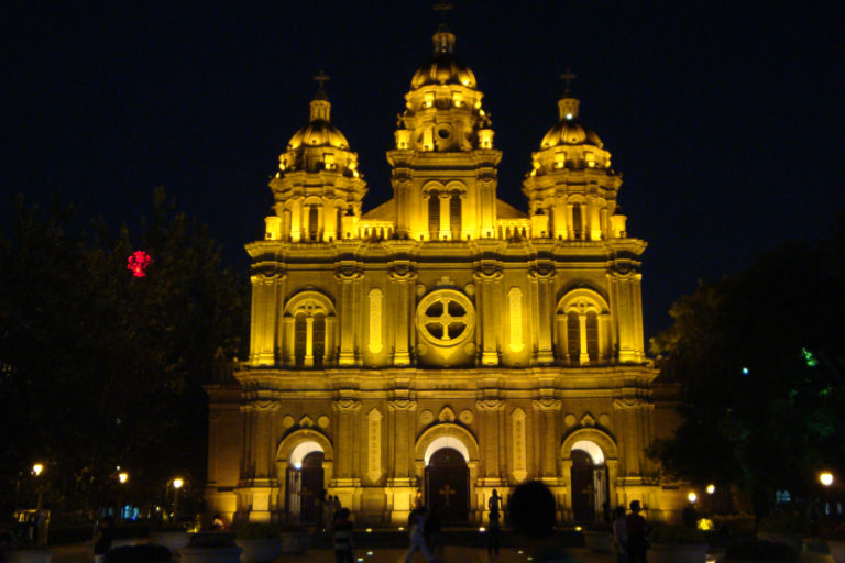Wide frontal night view of the illuminated three-towered facade of St. Joseph's Church on Wangfujing in Beijing, with trees and visitors in the dark foreground.