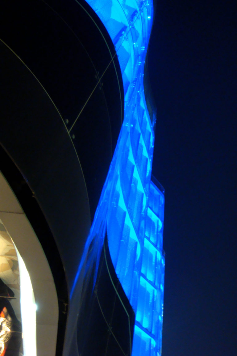 Nighttime close-up of an electric-blue illuminated tensile membrane canopy at a modern commercial complex in Beijing, framed by dark curved building surfaces.