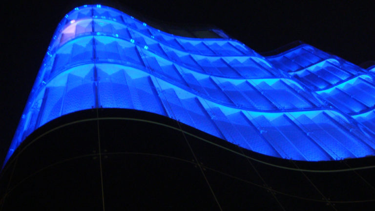 Low-angle nighttime view of a blue-lit translucent tensile canopy framed by dark curved building surfaces in a Beijing commercial complex.