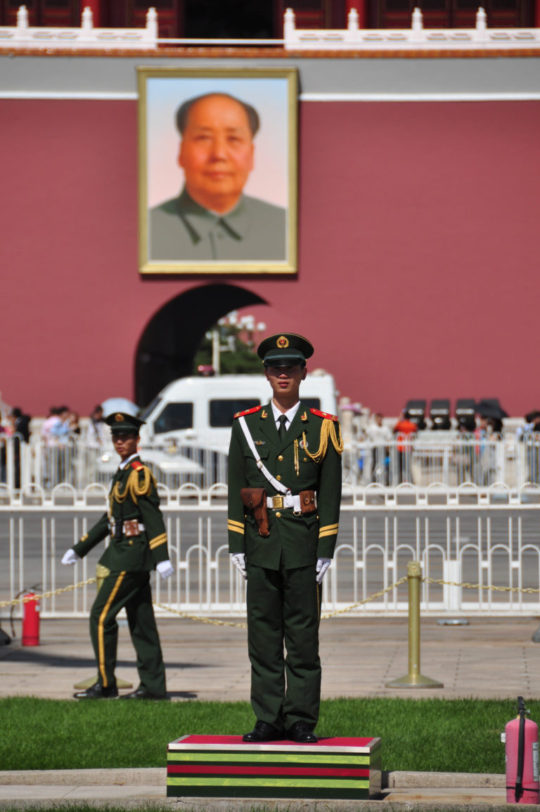 Two Chinese ceremonial guards in dark green uniforms near Tiananmen Gate, one standing frontally and one walking behind a barrier.