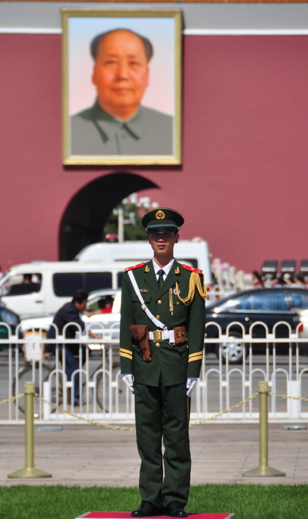 Chinese ceremonial guard in dark green uniform standing at attention in front of the red wall and arched gateway of Tiananmen Gate.