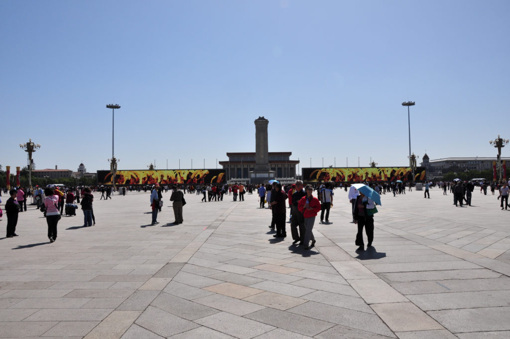Panoramic view across the wide paved expanse of Tiananmen Square with the Monument to the People’s Heroes in the distance and visitors scattered across the square.