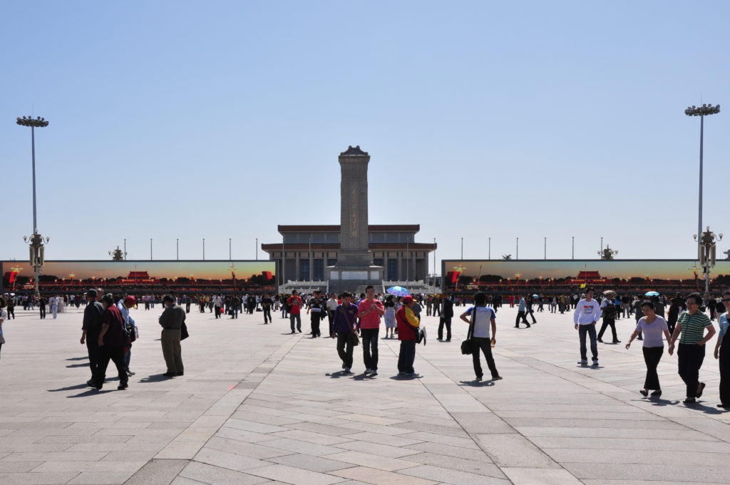 View across Tiananmen Square toward the Monument to the People’s Heroes and Chairman Mao Memorial Hall with crowds walking across the paved square on a clear day.