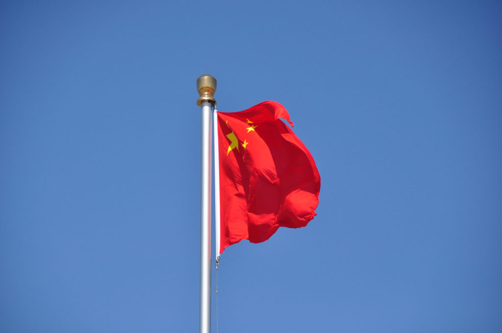 Flag of the People’s Republic of China flying from a tall white pole against a clear sky over Tiananmen Square.