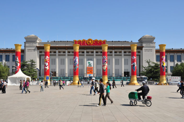 Great Hall of the People with a long colonnade and tall red-and-gold ceremonial columns set up in front for a national celebration.