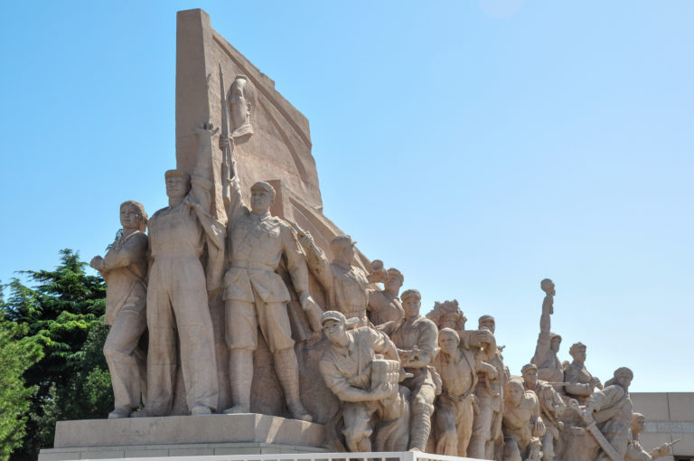 Side view of a long stone revolutionary sculpture showing idealized soldiers and civilians ascending toward a tall central slab against a clear blue sky.