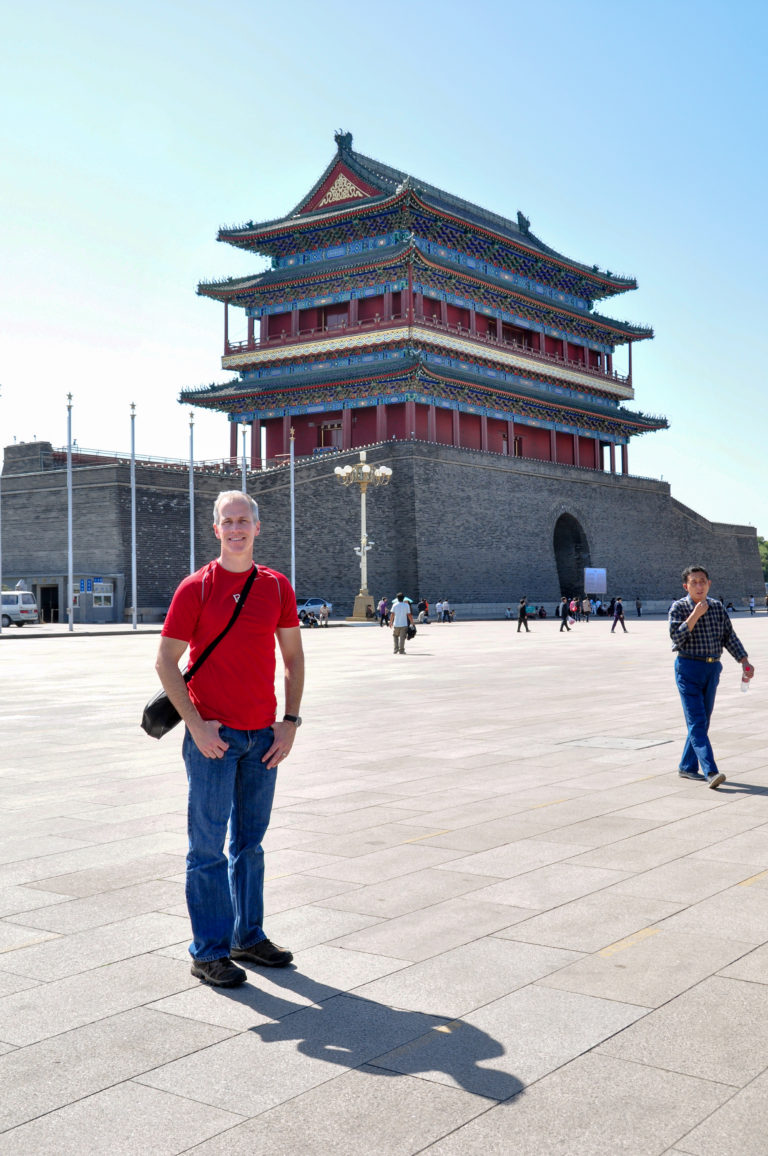 Man in a red shirt posing in the open square in front of Zhengyangmen Arrow Tower with its gray base and red upper structure.