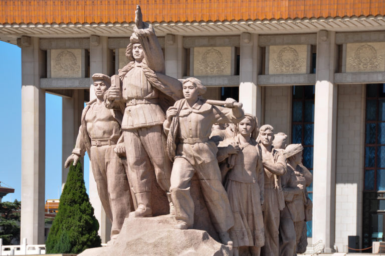 Stone revolutionary figural group showing a central man in military-style clothing with workers and women in front of a large civic building facade.