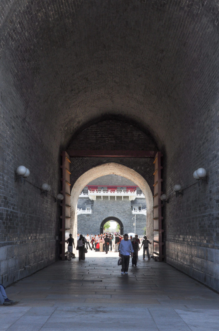 Axial view through nested gray-brick archways in Zhengyangmen, framing a distant gateway with small visitors walking through the passage.