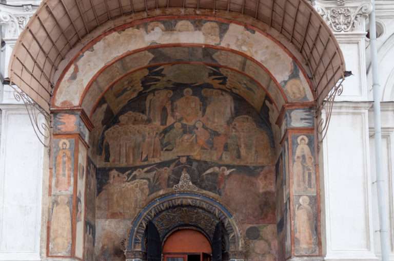 Weathered religious frescoes surrounding an arched doorway in the white exterior wall of the Assumption Cathedral.