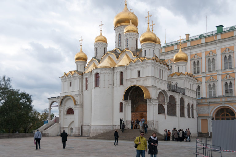 White Cathedral of the Annunciation with gilded domes and arched porch, with visitors standing in Cathedral Square.