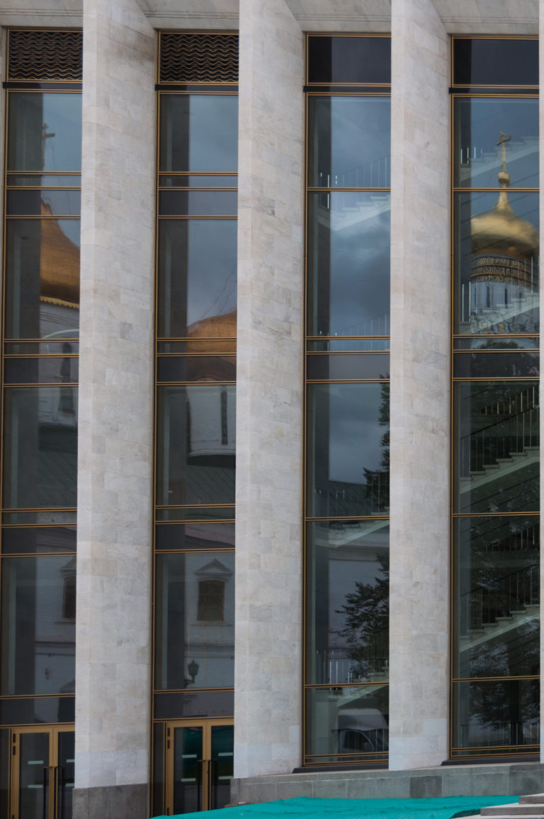 Tall glass bays and pale stone piers of the State Kremlin Palace reflecting a gilded dome and historic Kremlin buildings.
