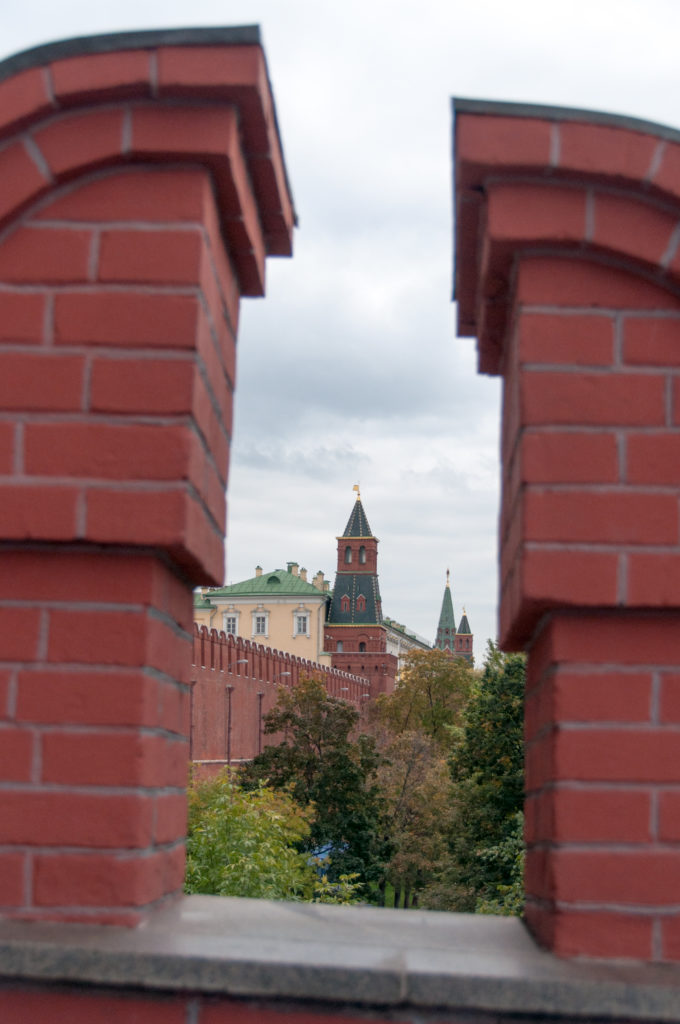 Distant Kremlin wall and roofed towers framed between two close red-brick parapets above autumn trees.