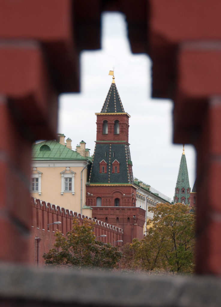 Kremlin tower and wall seen through a narrow opening in red-brick defensive masonry, with pale buildings beyond.