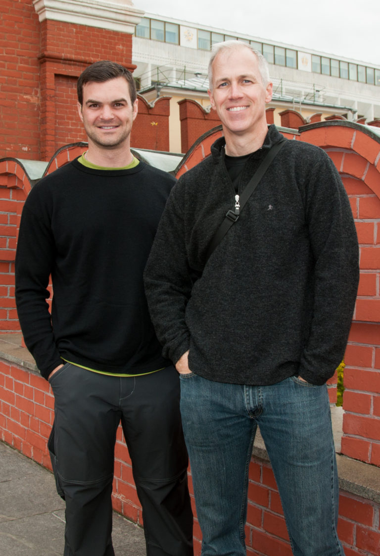 Two visitors standing side by side in front of a red-brick parapet and pale stone architecture.