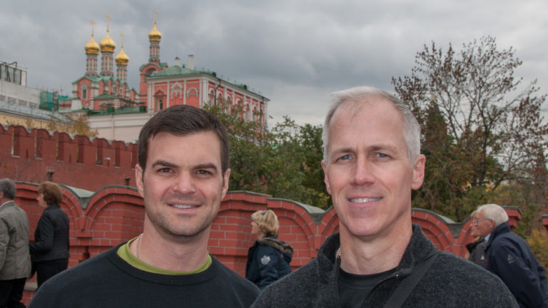 Two visitors posing near red Kremlin walls and colorful historic architecture, with other pedestrians behind them.