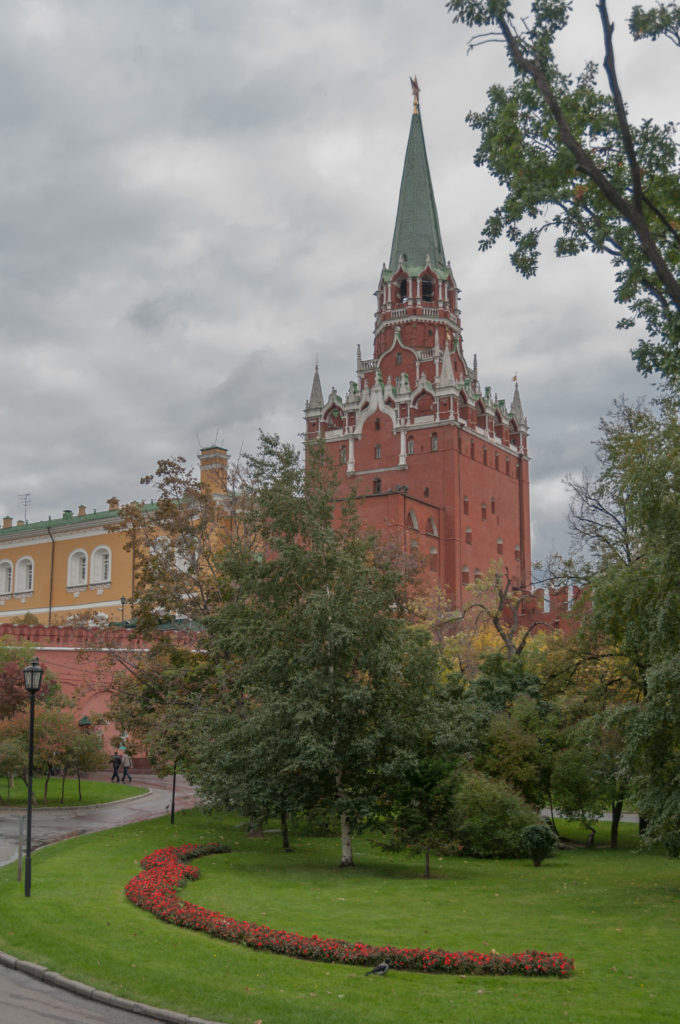 Star-topped red-brick Kremlin tower with green roof beyond lawns, flower beds, shrubs, and autumn trees.