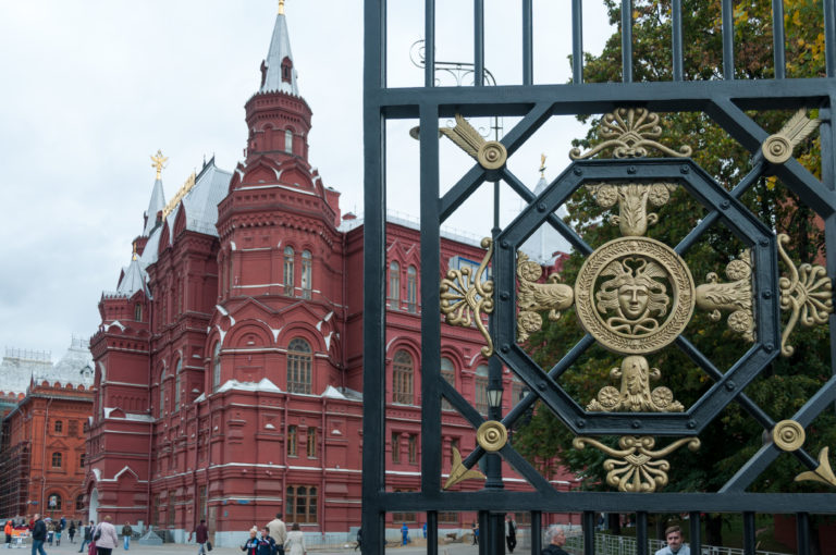 Ornate black-and-gold metal gate in the foreground with the red-brick State Historical Museum rising behind on Red Square.