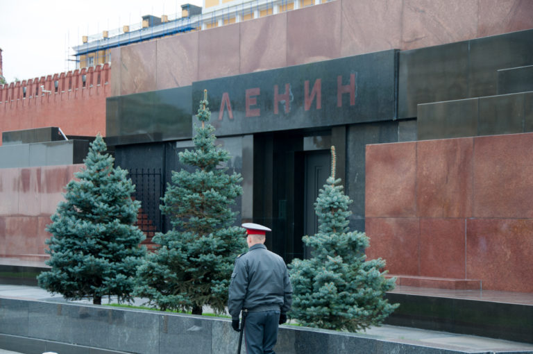 Uniformed guard standing near evergreen trees outside the dark entrance to Lenin Mausoleum, beneath the red Cyrillic inscription.