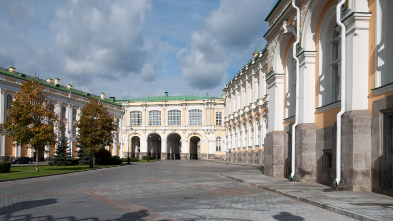 Paved Kremlin courtyard bordered by pale yellow-and-white palace buildings with tall arched windows and a three-arched passage.
