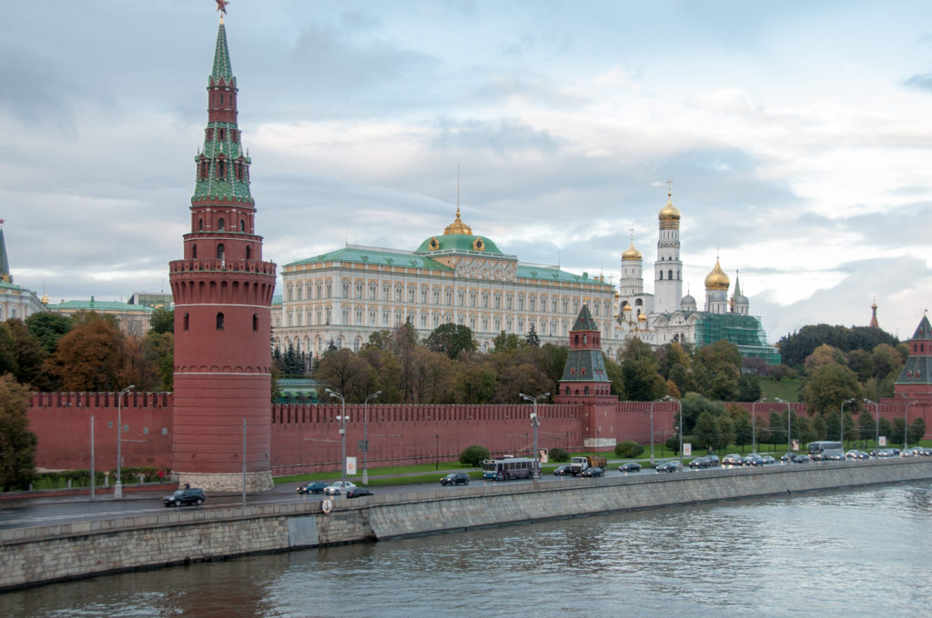Kremlin tower and red wall in front of the Grand Kremlin Palace, with white bell towers and golden domes to the right.
