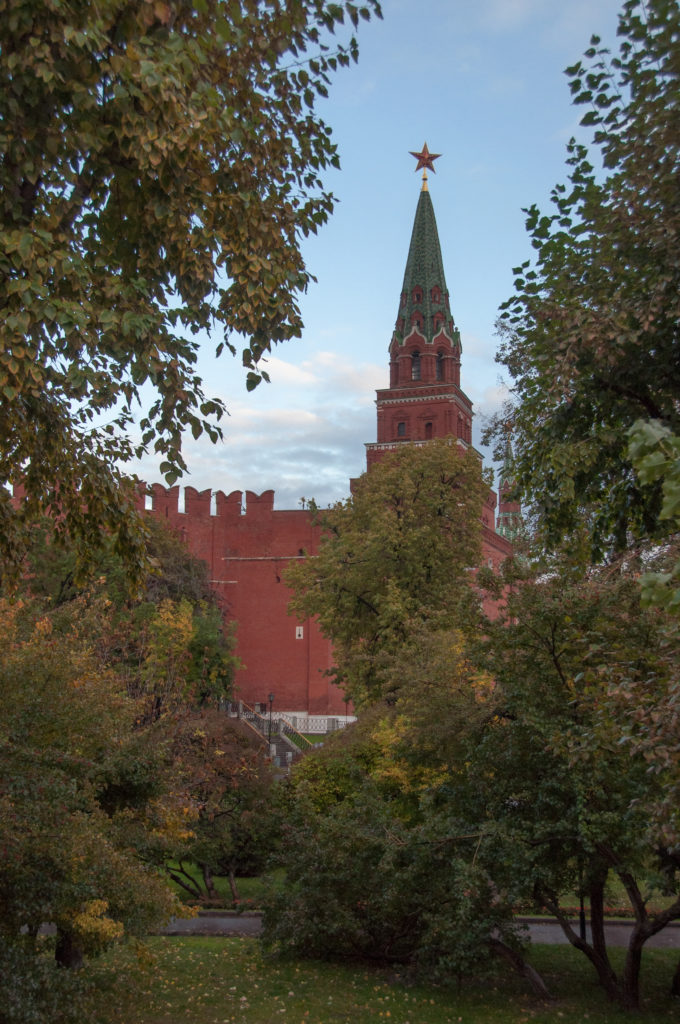 Red-brick Kremlin tower with green tent roof and red star above the wall, framed by trees in a garden.