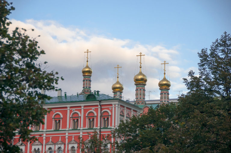 Red-and-white Orthodox church with four gilded onion domes rising behind leafy trees near the Kremlin.