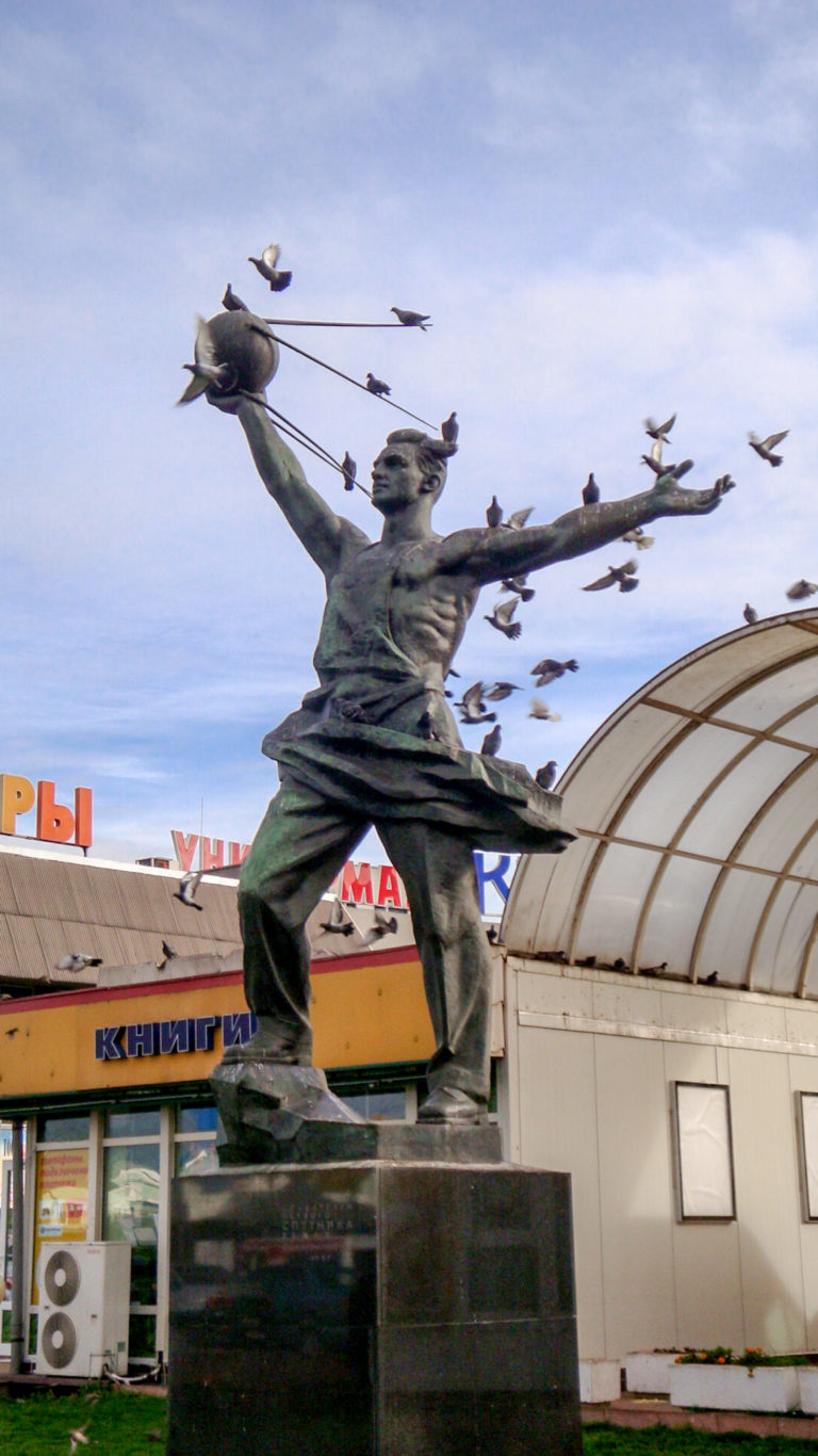 Bronze striding man raising an aircraft-shaped object, with additional airplane forms behind and a low building in the background.