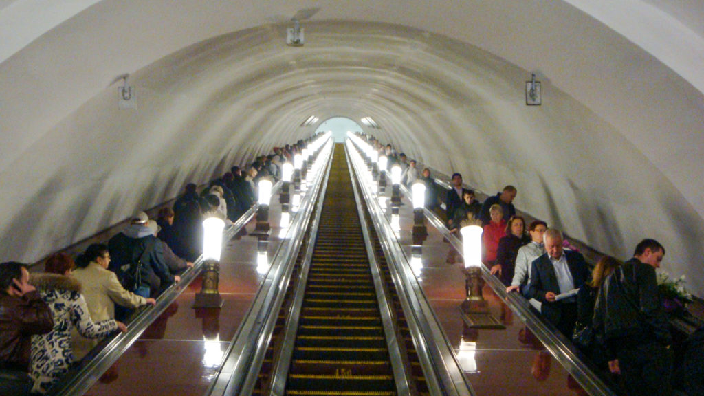 Crowded escalators rising through a white arched shaft toward a bright opening, divided by rows of glowing lamps.