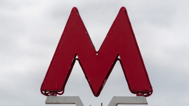 Large red angular Moscow Metro M entrance sign silhouetted against a pale daytime sky.