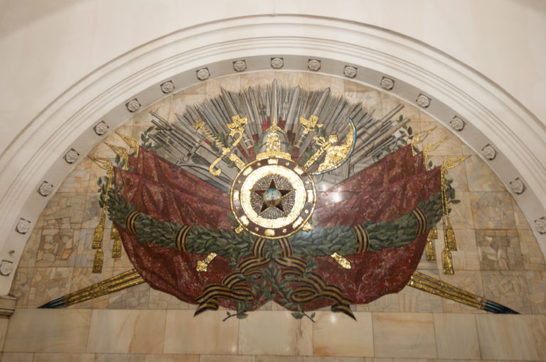 Large semicircular Soviet mosaic with central star, red banners, gold rays, foliage, and military motifs beneath an arch.