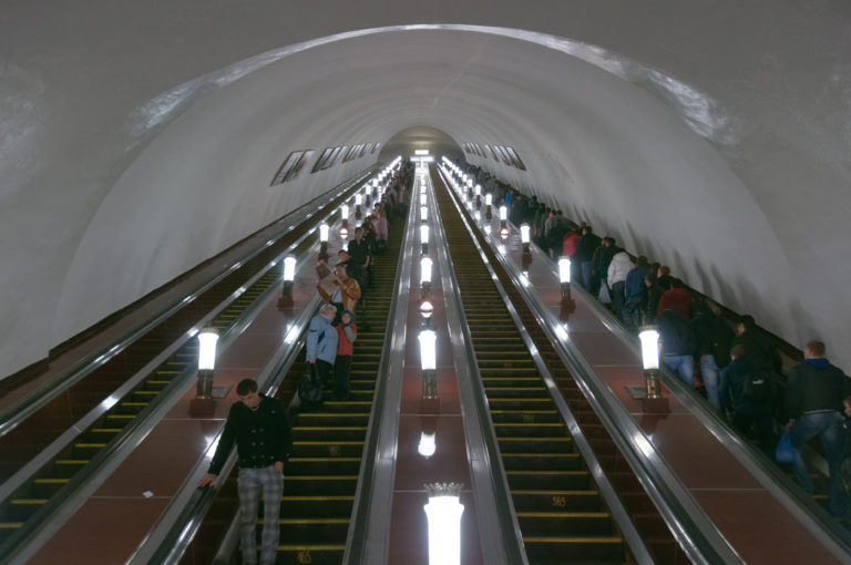 Symmetrical escalators climbing through a pale arched shaft toward a bright landing, lined with lamps and passengers.