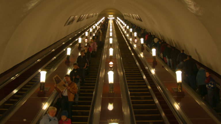 Long illuminated metro escalators rising steeply through an arched tunnel, with passengers traveling in both directions.