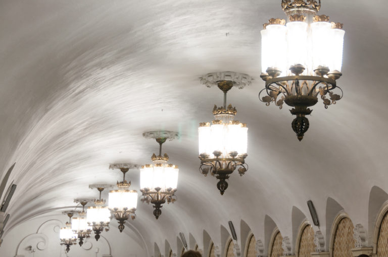 Row of ornate chandeliers with bright glass shades receding along a white vaulted metro ceiling above arched wall bays.