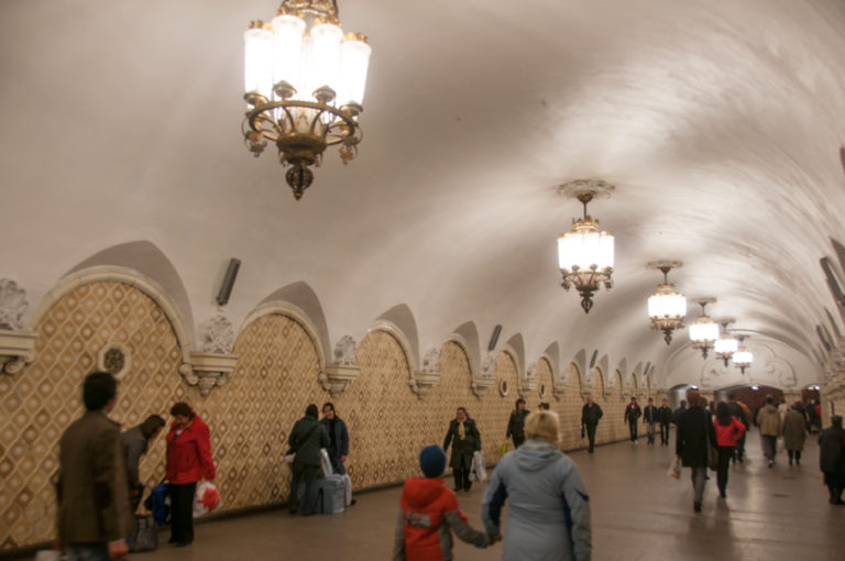 Passengers walking through a white-vaulted metro corridor with ornate chandeliers and repeating patterned arched wall bays.